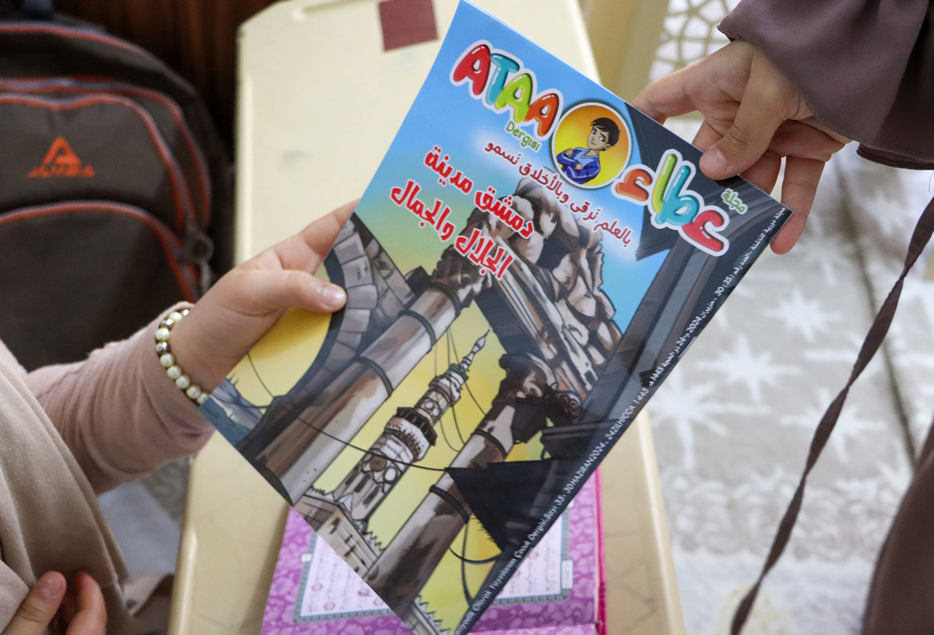 يد طفلة تستلم نسخة من مجلة عطاء من يد المعلمة في مركز تحفيظ القرآن الكريم